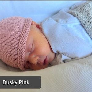 May include: A sleeping baby wearing a dusky pink knitted hat with a small ear detail. The baby is lying on a white surface, covered with a white onesie and a light grey blanket.