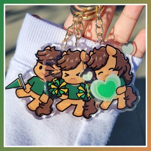 May include: A clear acrylic keychain featuring three cartoon pony characters with brown hair and tails, wearing green shirts. One holds a green flag, another pom-poms, and the last a green heart. The keychain has a gold chain.