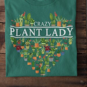 Puede incluir: Una camiseta verde con un dise&ntilde;o blanco en forma de coraz&oacute;n que presenta una variedad de plantas en macetas. El texto "CRAZY PLANT LADY" est&aacute; escrito en blanco sobre el coraz&oacute;n.