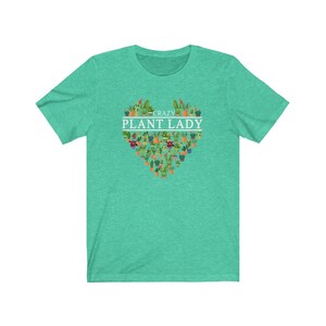 Puede incluir: Una camiseta verde turquesa con un dise&ntilde;o en forma de coraz&oacute;n blanco con el texto "Crazy Plant Lady" rodeado de una variedad de plantas en macetas.
