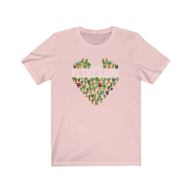 Puede incluir: Camiseta rosa de manga corta con un dise&ntilde;o blanco en forma de coraz&oacute;n que presenta una variedad de plantas en macetas y el texto "Crazy Plant Lady".
