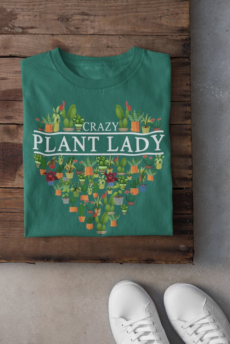Puede incluir: Una camiseta verde con un dise&ntilde;o blanco en forma de coraz&oacute;n que presenta una variedad de plantas en macetas. El texto "CRAZY PLANT LADY" est&aacute; escrito en blanco sobre el coraz&oacute;n.