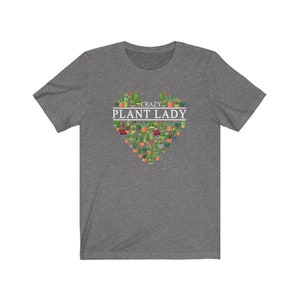 Puede incluir: Una camiseta gris jaspeada con un dise&ntilde;o en forma de coraz&oacute;n hecho de plantas en macetas. El texto "Crazy Plant Lady" est&aacute; impreso en blanco sobre el coraz&oacute;n.