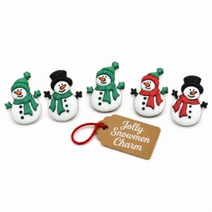 May include: Five white snowman-shaped buttons with orange carrot noses and black button details. Three snowmen wear green scarves and hats, while two wear red scarves and black top hats. A brown rectangular tag with the text Jolly Snowmen Charm.
