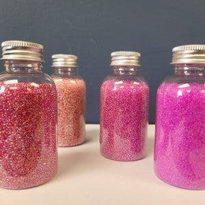 May include: Four clear glass bottles filled with iridescent pink, light pink, and magenta beads. The bottles have silver lids.