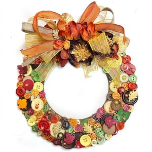 May include: A circular wreath covered in assorted buttons in shades of red, orange, yellow, green, and brown. The wreath features decorative leaf-shaped buttons and a large bow at the top made of orange, yellow, and brown ribbons and fabric flowers.