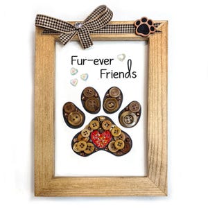 May include: A framed artwork with a paw print design made of brown buttons. The artwork includes the text "Fur-ever Friends" and is decorated with a checkered bow and a paw print accent. The frame is made of light-colored wood.