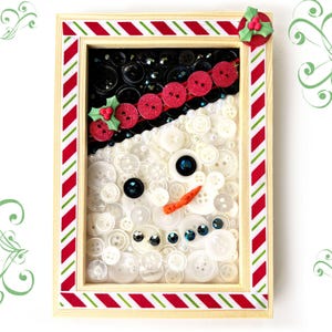 May include: A framed snowman art piece made from buttons. The snowman's hat is black buttons, the body is white buttons, and the face has a carrot nose and black and blue button eyes. The frame is decorated with red and green striped ribbon.