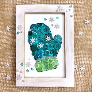 May include: A framed artwork featuring a mitten design crafted from various shades of green and teal buttons. The mitten is set against a white background, framed in a distressed white wooden frame, and adorned with snowflake and pearl embellishments.