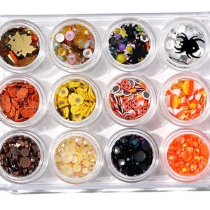 May include: A clear plastic container with 12 small jars filled with various Halloween-themed nail art embellishments. The jars contain orange, black, white, yellow, and brown glitter, as well as small plastic pumpkins, candy corn, and spiders.