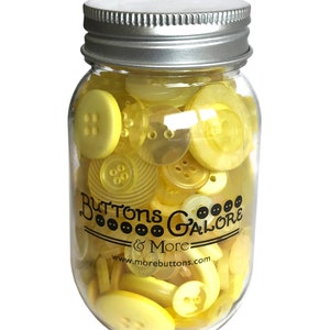 May include: A glass jar filled with yellow buttons of various sizes and shapes. The jar has a silver lid and a black label that reads "Buttons Galore & More" with the website address "www.morebuttons.com".