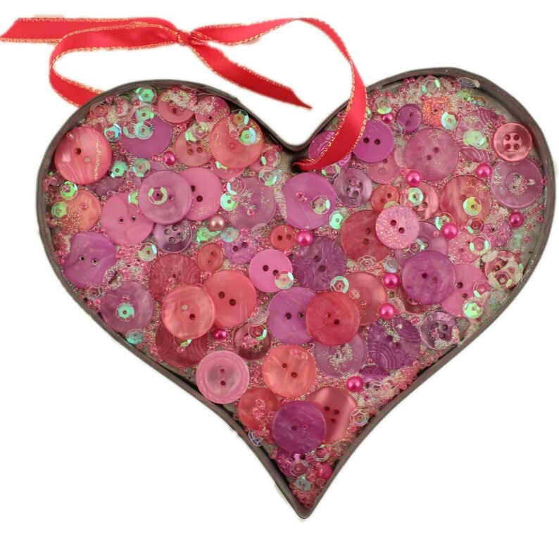 May include: A heart-shaped metal frame filled with pink, red, and white buttons and sequins. The frame has a red ribbon attached to the top.