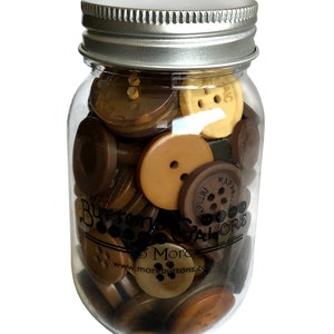May include: A clear glass jar filled with various brown buttons. The buttons are in shades of tan, light brown, and dark brown. The jar has a silver lid and the text "Buttons Galore" is printed on the jar.