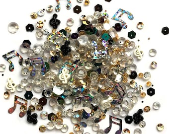 Op de afbeelding: Een mix van iriserende en goudkleurige sequins, strass steentjes en muzieknoot bedels. De bedels zijn in zwart, wit en iriserende kleuren.