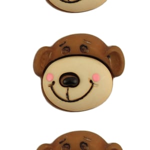 May include: Three brown and beige plastic buttons shaped like monkey faces with pink cheeks and black eyes.