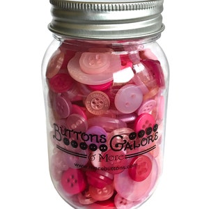 May include: A glass jar filled with pink, red, and clear buttons. The jar has a silver lid and a black label that reads "Buttons Galore & More" with the website address "www.morebuttons.com".