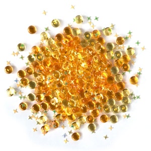 Op de afbeelding: Een hoop kleine, ronde, doorschijnende amberkleurige kralen met gouden glittersterren eromheen gestrooid.