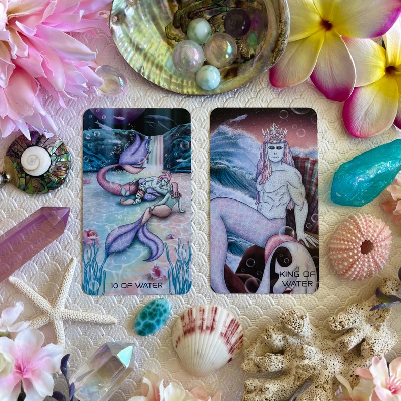 Puede incluir: Dos cartas de tarot, el 10 de Agua y el Rey de Agua, se muestran sobre una superficie blanca rodeada de conchas marinas, flores y cristales. La carta del 10 de Agua representa a dos sirenas con colas moradas, mientras que la carta del Rey de Agua muestra a un trit&oacute;n con una corona y una cola morada.