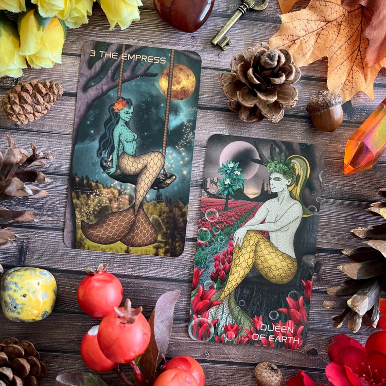 Puede incluir: Dos cartas de tarot, "3 La Emperatriz" y "Reina de la Tierra", con ilustraciones de sirenas. La carta de la Emperatriz muestra una sirena de piel verde con una cola marr&oacute;n sentada en una rama de &aacute;rbol. La carta de la Reina de la Tierra muestra una sirena con una cola amarilla sentada en un toc&oacute;n de &aacute;rbol en un campo de flores rojas.