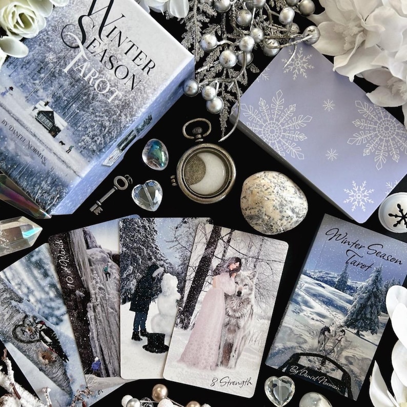 Peut inclure: Un jeu de tarot "Winter Season" avec une bo&icirc;te bleue et blanche repr&eacute;sentant un paysage enneig&eacute;. La bo&icirc;te porte l'inscription "Winter Season Tarot" sur le devant. Le jeu est dispos&eacute; sur une surface noire avec d'autres cartes de tarot, des cristaux et une montre de poche argent&eacute;e.