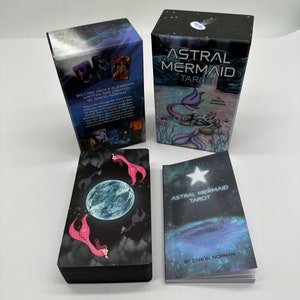 Puede incluir: Un juego de cartas de Tarot Astral Mermaid con un libro de gu&iacute;a y una caja. La caja es negra con un fondo de cielo estrellado y presenta una sirena con una cola azul y una corona. El libro de gu&iacute;a es azul con una estrella y el texto "Astral Mermaid Tarot" y "Por Daniel Norman". Las cartas son negras con una luna azul y sirenas con colas rosas.