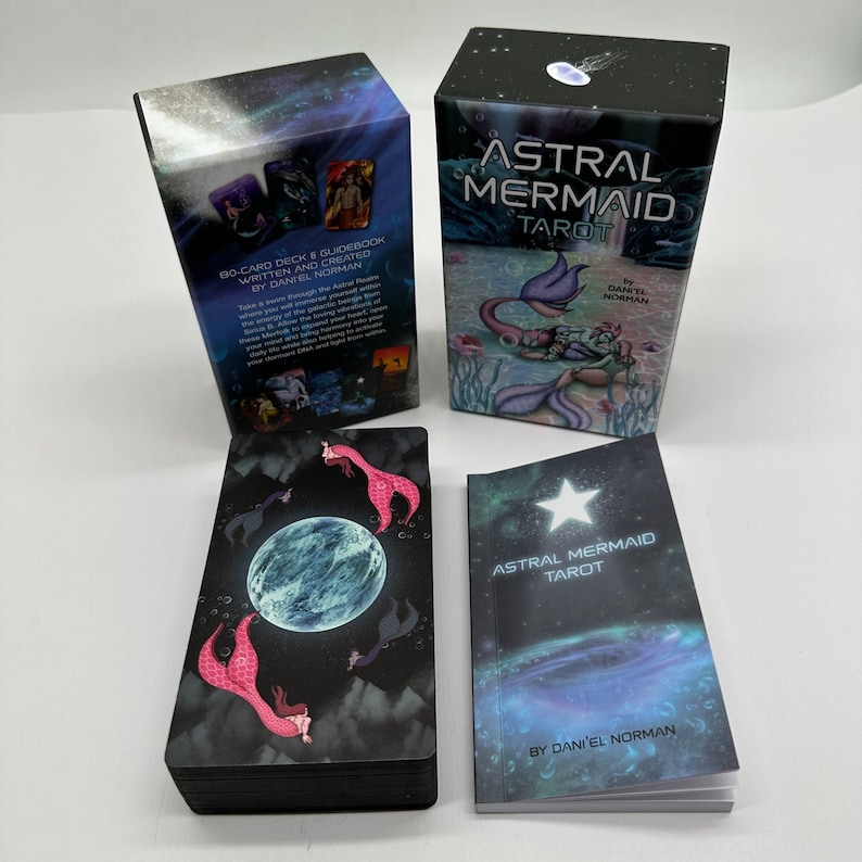 Puede incluir: Un juego de cartas de Tarot Astral Mermaid con un libro de gu&iacute;a y una caja. La caja es negra con un fondo de cielo estrellado y presenta una sirena con una cola azul y una corona. El libro de gu&iacute;a es azul con una estrella y el texto "Astral Mermaid Tarot" y "Por Daniel Norman". Las cartas son negras con una luna azul y sirenas con colas rosas.