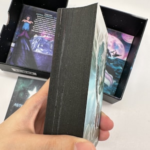 Puede incluir: Una baraja de cartas de Tarot Astral Mermaid con un dorso negro y una imagen colorida de una sirena en el frente. Las cartas est&aacute;n en una caja negra con un cielo morado y rosa y una ballena blanca.