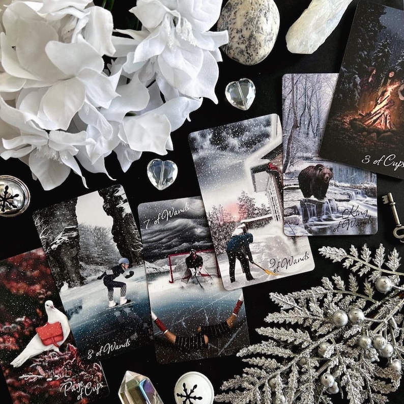 Peut inclure: Un tirage de cartes de tarot avec dix cartes dispos&eacute;es sur une surface noire. Les cartes sont du jeu de tarot Wild Unknown et pr&eacute;sentent des sc&egrave;nes hivernales. Les cartes sont&nbsp;: Page de Coupes, 8 de B&acirc;tons, 7 de B&acirc;tons, 2 de B&acirc;tons, Roi de B&acirc;tons, 3 de Coupes, et quelques autres cartes qui ne sont pas enti&egrave;rement visibles. Il y a aussi des fleurs blanches, des cristaux et d'autres objets d&eacute;coratifs sur la surface.