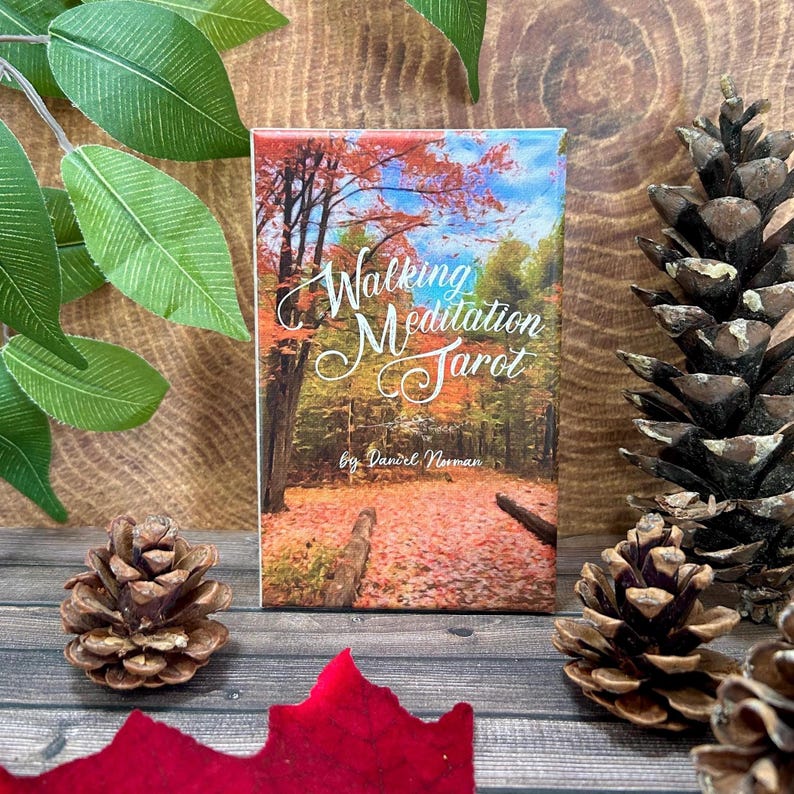 May include: A box for a deck of "Walking Meditation Tarot" cards. The box features a photograph of a forest path in autumn with red, orange, and yellow leaves. The text on the box reads "Walking Meditation Tarot" by "Daniel Norman".