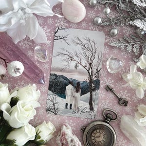 Peut inclure: Une carte de tarot repr&eacute;sentant une sc&egrave;ne hivernale avec deux personnages debout dans la neige. La carte est intitul&eacute;e "Les Amoureux" et est entour&eacute;e de fleurs blanches, de cristaux et d'une montre de poche argent&eacute;e.