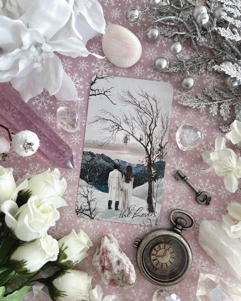Peut inclure: Une carte de tarot repr&eacute;sentant une sc&egrave;ne hivernale avec deux personnages debout dans la neige. La carte est intitul&eacute;e "Les Amoureux" et est entour&eacute;e de fleurs blanches, de cristaux et d'une montre de poche argent&eacute;e.