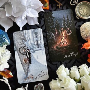 Peut inclure: Deux cartes de tarot, 3 d'&Eacute;p&eacute;es et 3 de Coupes, avec des sc&egrave;nes hivernales. La carte 3 d'&Eacute;p&eacute;es montre un pic sur un tronc d'arbre dans une for&ecirc;t enneig&eacute;e. La carte 3 de Coupes montre trois personnages r&eacute;unis autour d'un feu de joie dans une for&ecirc;t enneig&eacute;e.