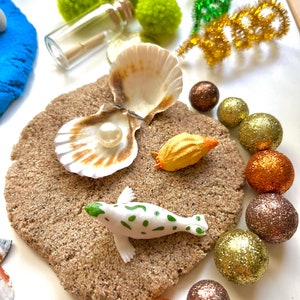 Pu&ograve; includere: Una statuina di foca bianca con macchie verdi su una superficie di sabbia marrone. Ci sono due conchiglie, una con una perla all'interno, e una piccola conchiglia arancione. Ci sono anche diverse sfere luccicanti dorate, marroni e arancioni.