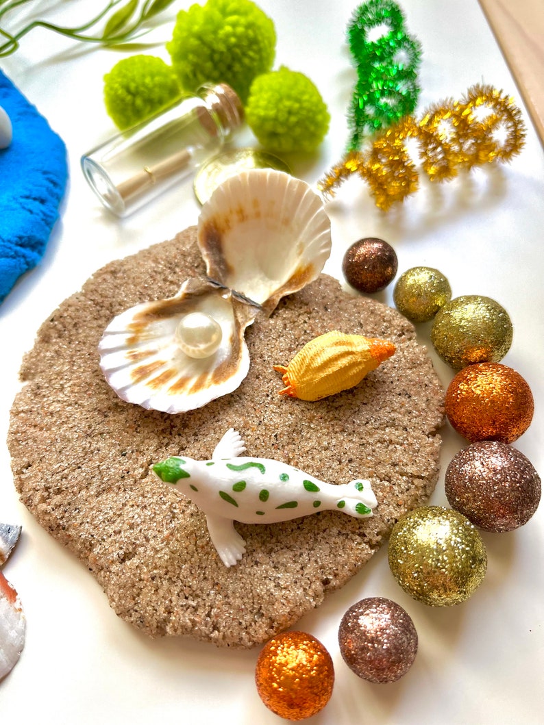Pu&ograve; includere: Una statuina di foca bianca con macchie verdi su una superficie di sabbia marrone. Ci sono due conchiglie, una con una perla all'interno, e una piccola conchiglia arancione. Ci sono anche diverse sfere luccicanti dorate, marroni e arancioni.
