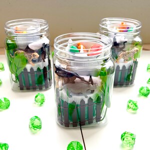 May include: Three clear glass jars filled with small plastic animal figurines, green plastic plants, and a white base with a black picket fence. The jars are on a white surface with green gem-like stones scattered around them.