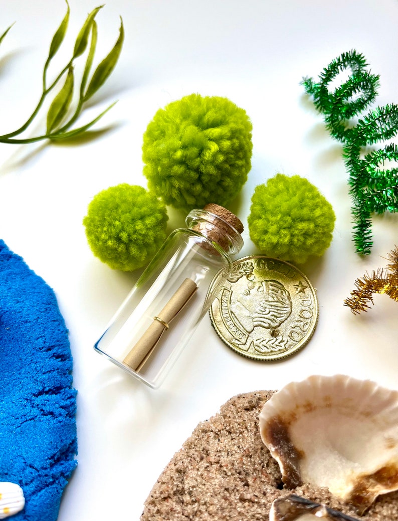 Pu&ograve; includere: Una bottiglia di vetro con un tappo di sughero che contiene un pezzo di carta arrotolato. La bottiglia &egrave; circondata da pompon verdi, una moneta d'oro e sabbia blu.