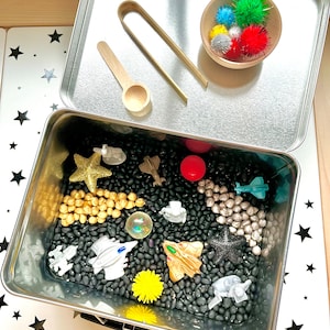 May include: A silver metal container with a black bean sensory bin. The bin contains small toy spaceships, a yellow star, a yellow flower, a clear plastic ball, and silver and gold beads. The lid of the container is open and shows a wooden spoon, a pair of metal tongs, and a small wooden bowl filled with colorful pom-poms.