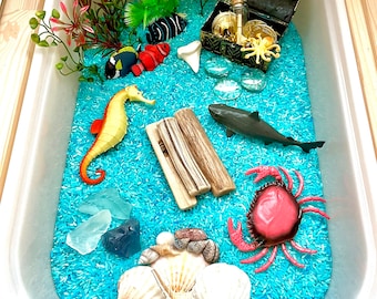 May include: A blue sensory bin filled with blue rice, featuring a variety of ocean-themed toys, including a shark, a crab, a seahorse, seashells, and a treasure chest with gold coins.