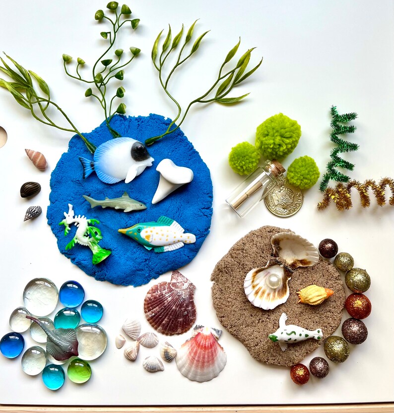 Pu&ograve; includere: Un vassoio di plastilina blu con una variet&agrave; di giocattoli marini, tra cui uno squalo, un cavalluccio marino, un pesce e una conchiglia. Ci sono anche alcune gemme di vetro e una piccola bottiglia con un tappo.