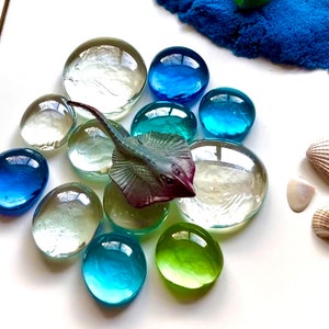 Pu&ograve; includere: Un mosaico di vetro blu e verde con una piccola statuina di razza di plastica al centro. Il mosaico &egrave; circondato da conchiglie bianche.