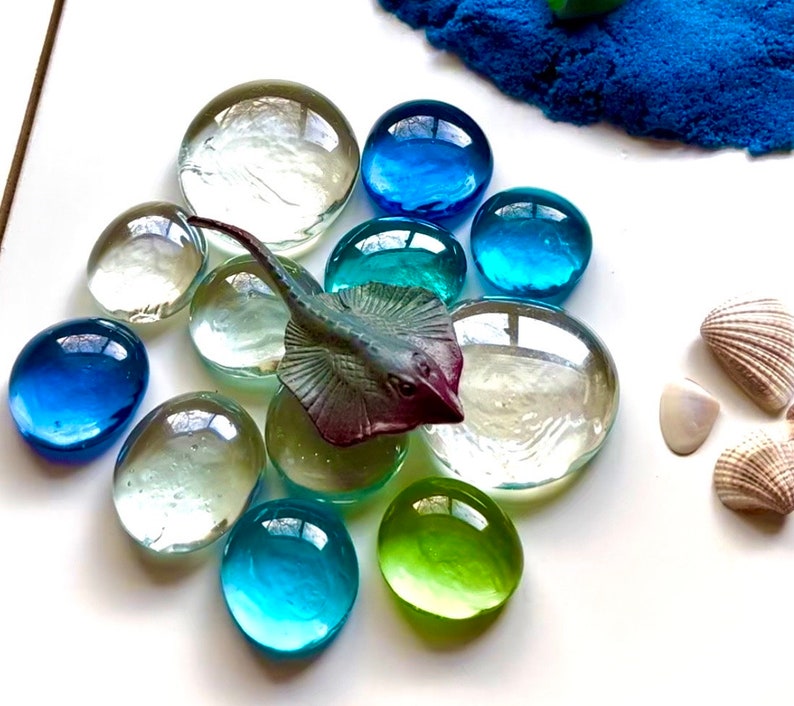 Pu&ograve; includere: Un mosaico di vetro blu e verde con una piccola statuina di razza di plastica al centro. Il mosaico &egrave; circondato da conchiglie bianche.