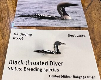 May include: A black and white enamel pin of a black-throated diver, a type of bird. The pin is labeled "UK Birding No. 96 Black-throated Diver Status: Breeding species Sept 2022 Limited Edition - Badge 51 of 150".