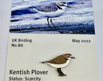 May include: A pin featuring a Kentish Plover bird design, with brown, white, and black enamel. The pin is a limited edition, badge 51 of 150. The image also includes a photo of the bird and text that reads "UK Birding No.86 May 2022".