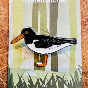 Könnte beinhalten: Metall-Anstecknadel mit einem Austernfischer-Vogel mit schwarzen, weißen und orangefarbenen Details. Die Anstecknadel befindet sich auf einer Karte mit dem Text "Oystercatcher" und "#11 Wild Britain" vor einem Waldbild.