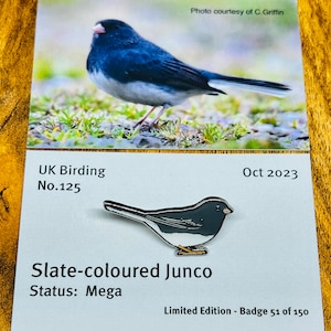 May include: A silver enamel pin of a slate-colored junco, a small songbird with a gray body, white belly, and black head. The pin is labeled "Slate-colored Junco" and "Status: Mega." The pin is part of a limited edition series, with the number "51 of 150" printed on the card.