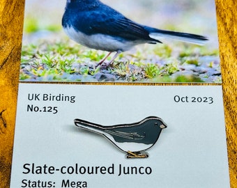 Op de afbeelding: Een zilveren emaille pin met een leisteen-junco, een kleine zangvogel met een grijs lichaam, witte buik en zwarte kop. De pin is gelabeld "Slate-colored Junco" en "Status: Mega." De pin is onderdeel van een limited edition serie, met het nummer "51 van 150" gedrukt op de kaart.