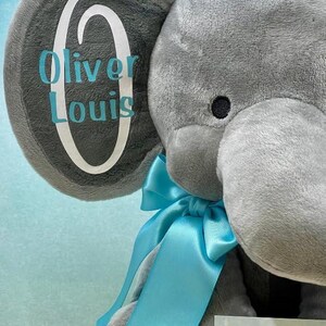 May include: A gray plush elephant with a blue ribbon and the name "Oliver Louis" on its ear. The elephant has a black eye and a tag that says "Creations by Emas".