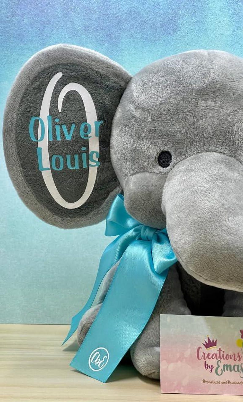 May include: A gray plush elephant with a blue ribbon and the name "Oliver Louis" on its ear. The elephant has a black eye and a tag that says "Creations by Emas".