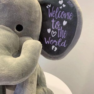 May include: A plush gray elephant toy with a dark gray ear. The ear features the text "Welcome to the World" in purple, along with hearts, a crown, footprints, and the year 2020. The elephant is soft and cuddly.