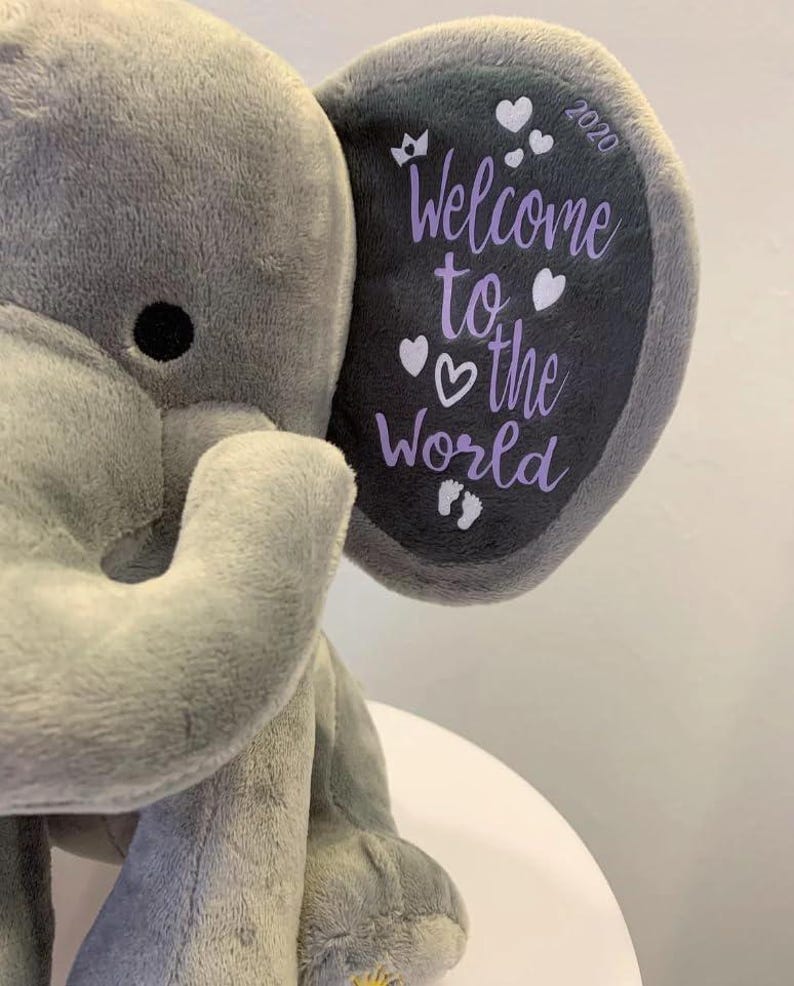 May include: A plush gray elephant toy with a dark gray ear. The ear features the text "Welcome to the World" in purple, along with hearts, a crown, footprints, and the year 2020. The elephant is soft and cuddly.